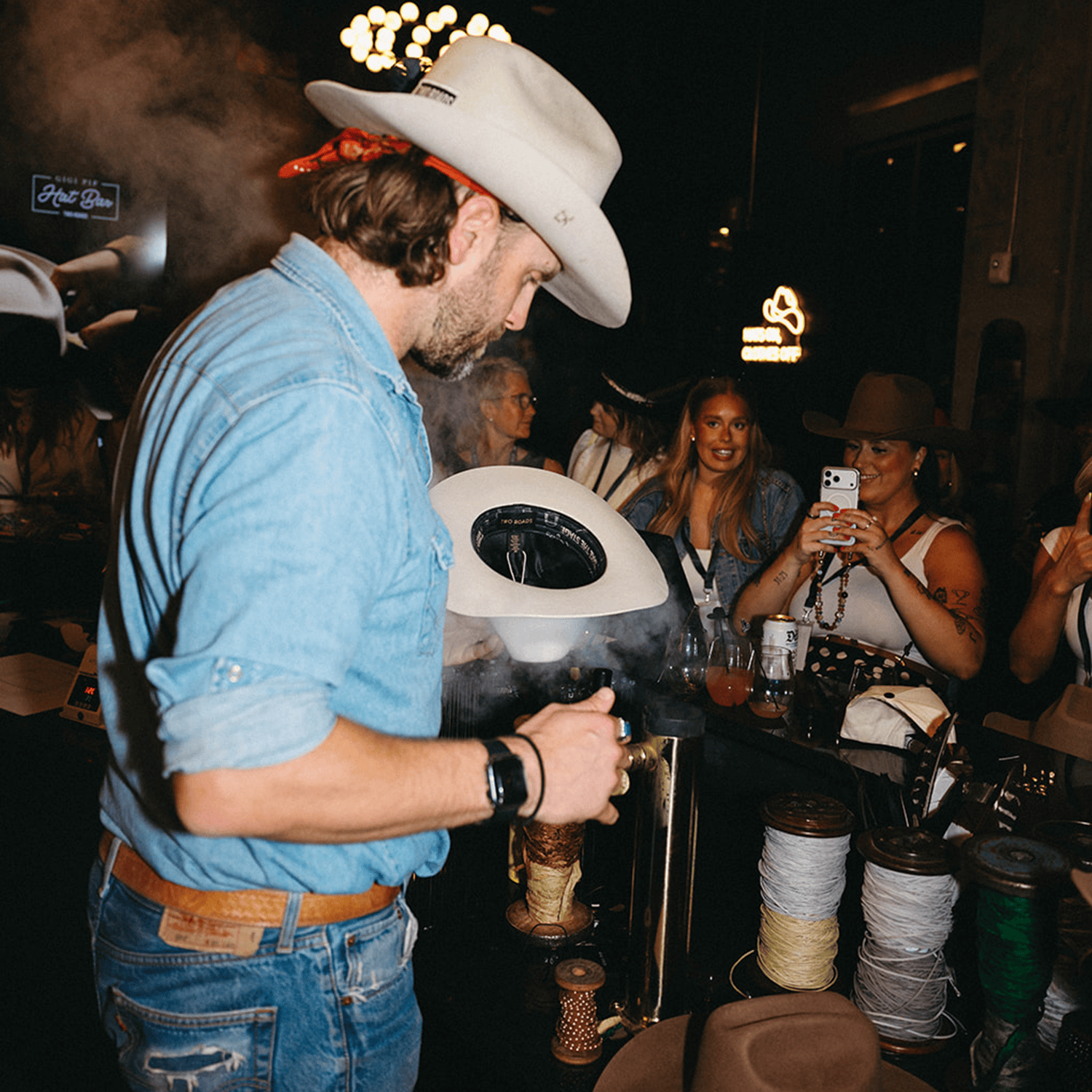Man in cowboy hat using a machine at a custom hat bar event with people in the background.