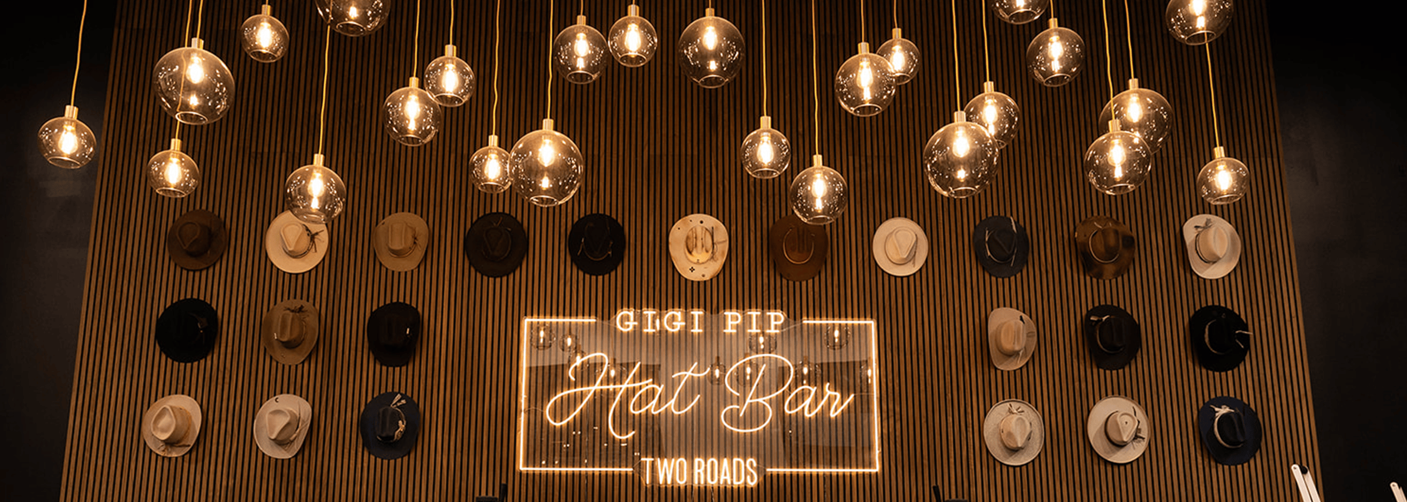 Decorative wall with hanging lights and a sign for 'Gigi Pip Hat Bar Two Roads' at a custom hat bar event.
