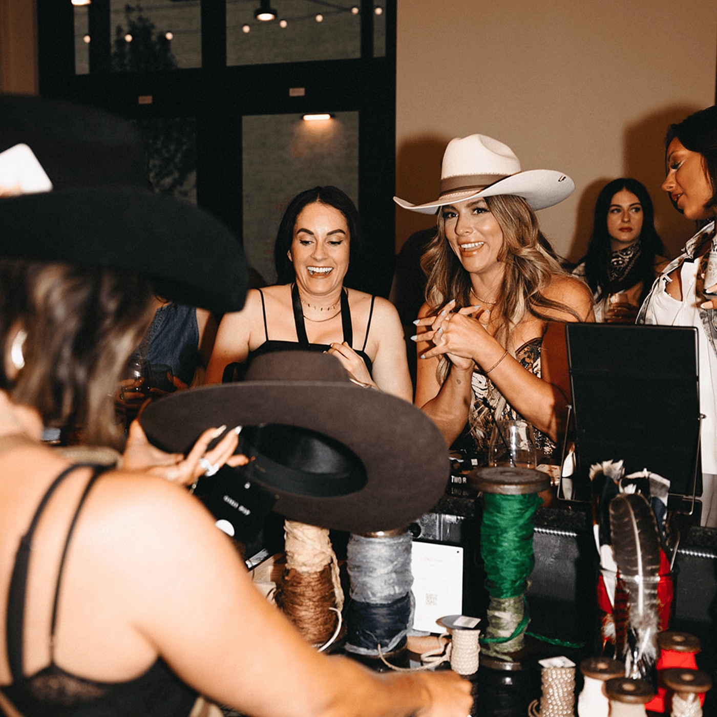 Group of women in a room with hats and yarn, possibly at a custom hat bar event.