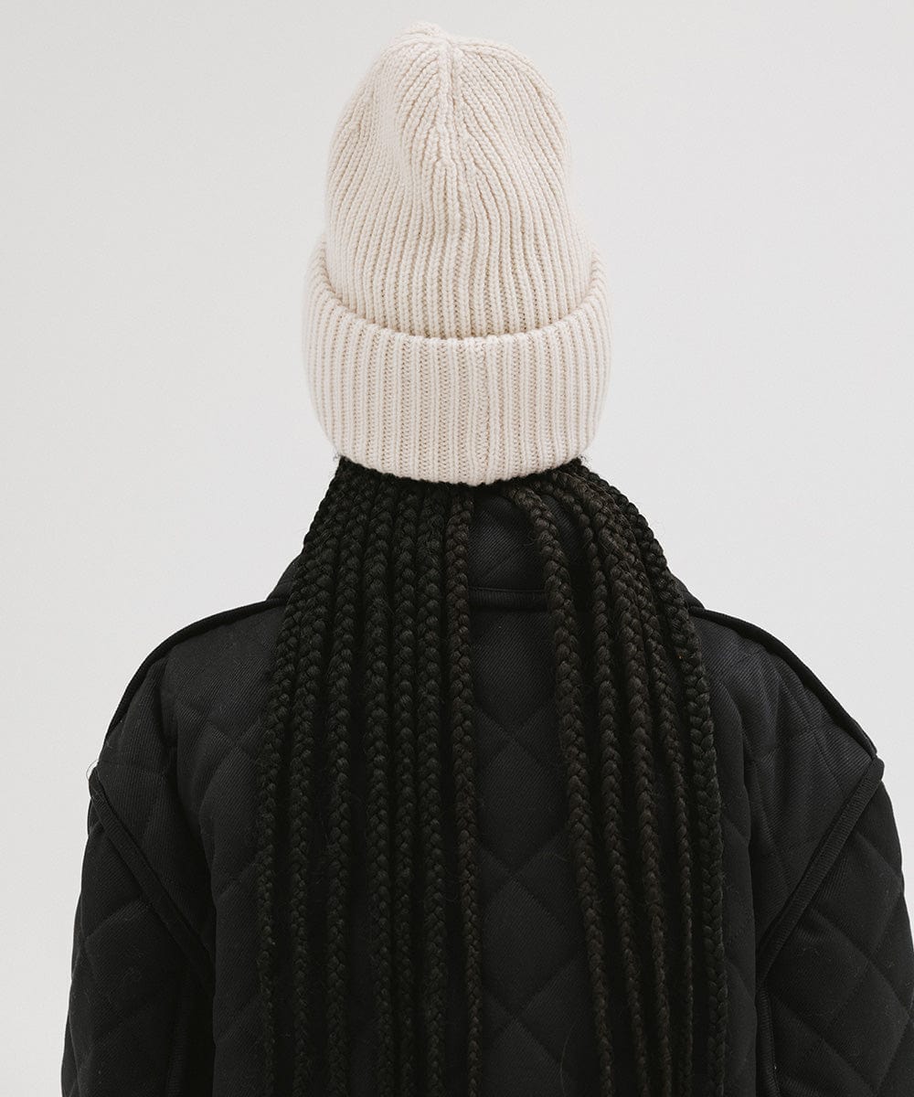 A person wearing an off white colored ribbed beanie and a black jacket, facing behind against a plain background #color_off white