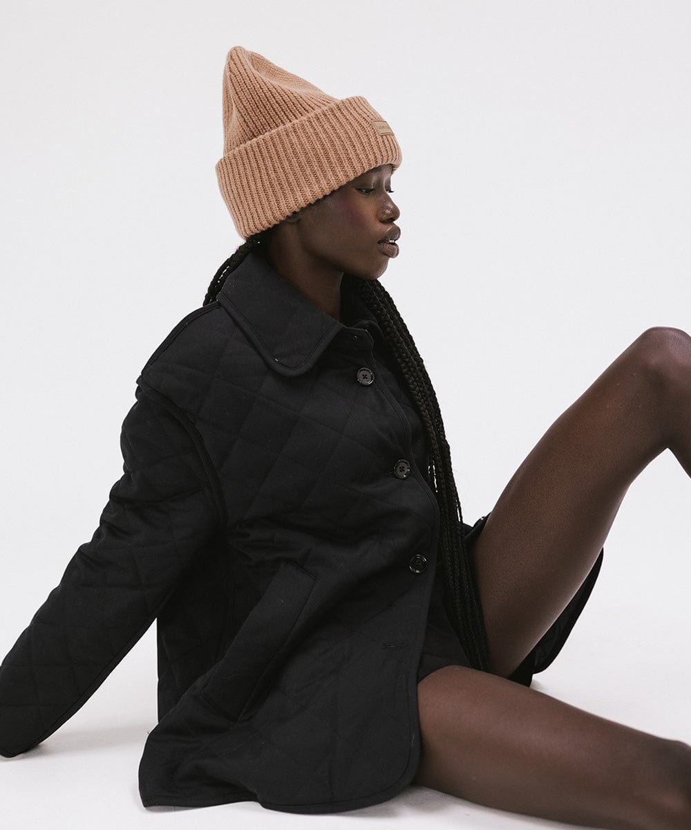 A woman wearing a brown colored ribbed beanie and a black jacket sitting on a plain background #color_brown