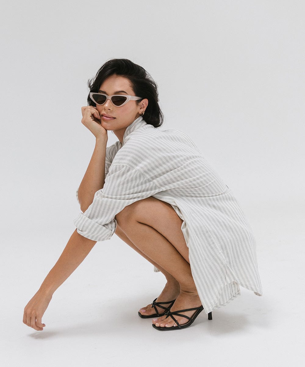 Woman wearing a light grey cat eye sunglasses, a white shirt with grey lines and black heels, sitting against a plain background #color_light grey
