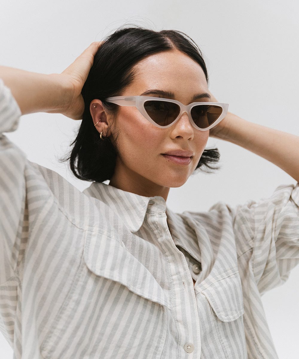 Woman wearing a light grey cat eye sunglasses and a white shirt with grey lines, looking sideways against a plain background #color_light grey