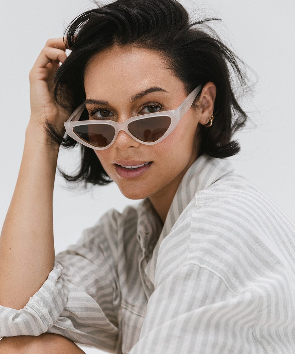 Woman wearing a light grey cat eye sunglasses and a white shirt with grey lines, posing against a plain background #color_light grey
