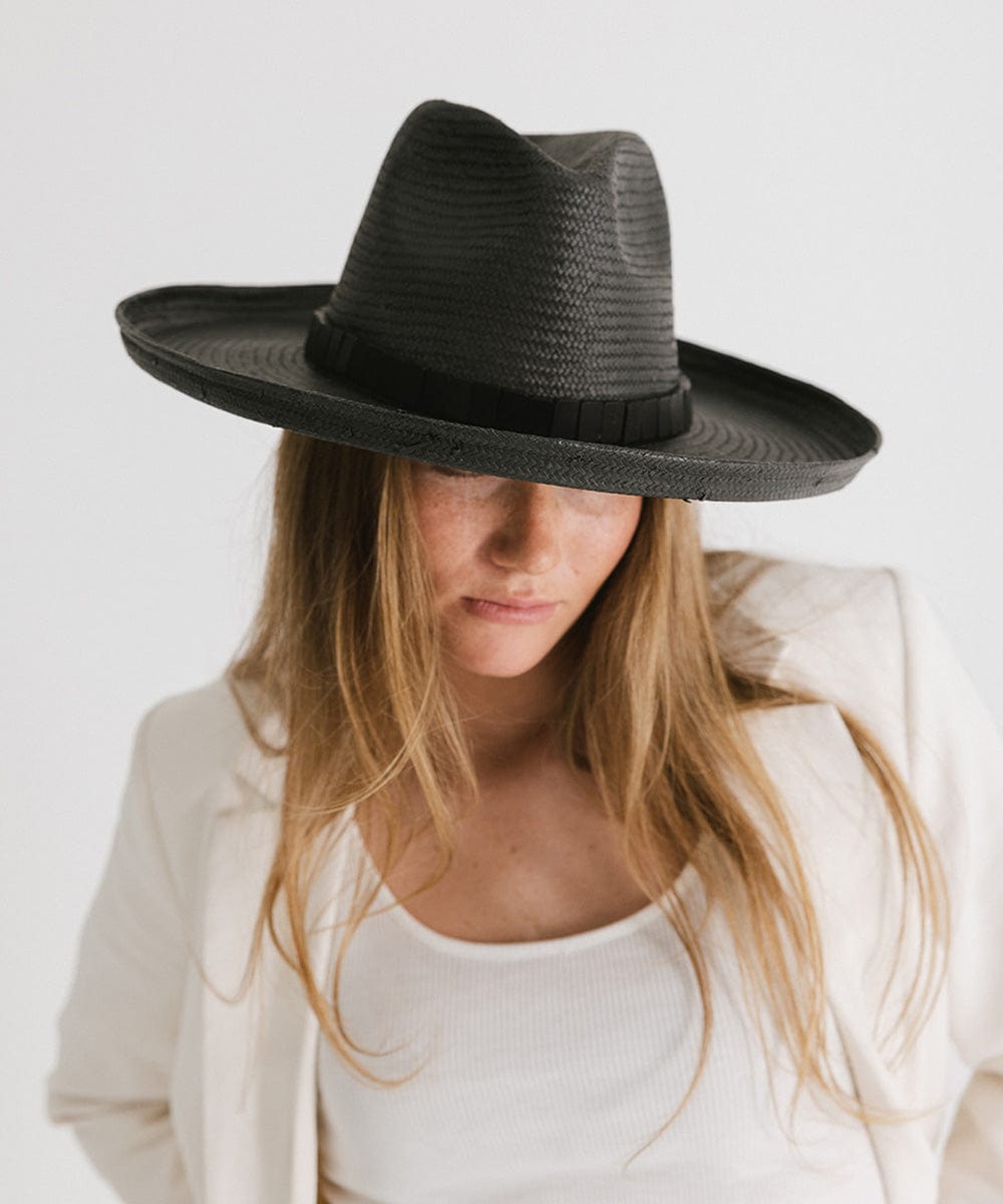 Woman wearing a black pencil brim straw hat with a hat band and a white top with a white blazer against a plain background #color_black