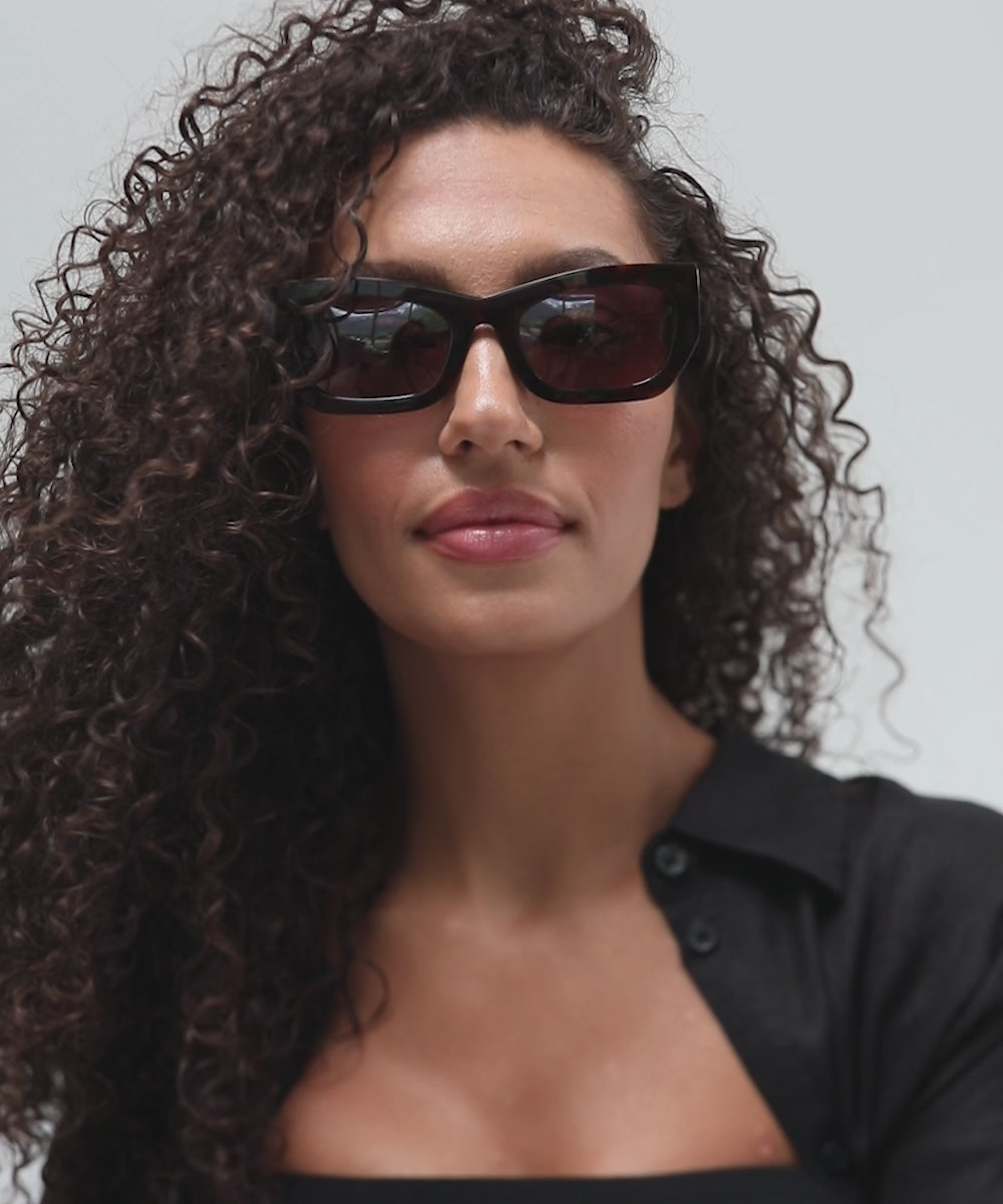 Woman wearing tortoise pattern sunglasses and a black shirt, posing against a plain background #color_tortoise