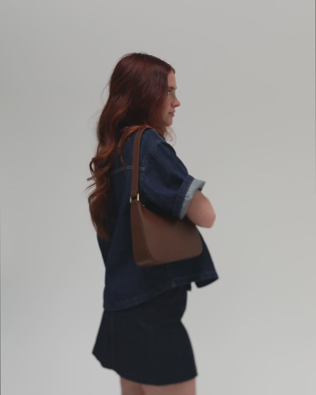 Woman wearing a denim shirt and a denim skirt, carrying a chocolate colored genuine leather handbag against a plain background #color_chocolate