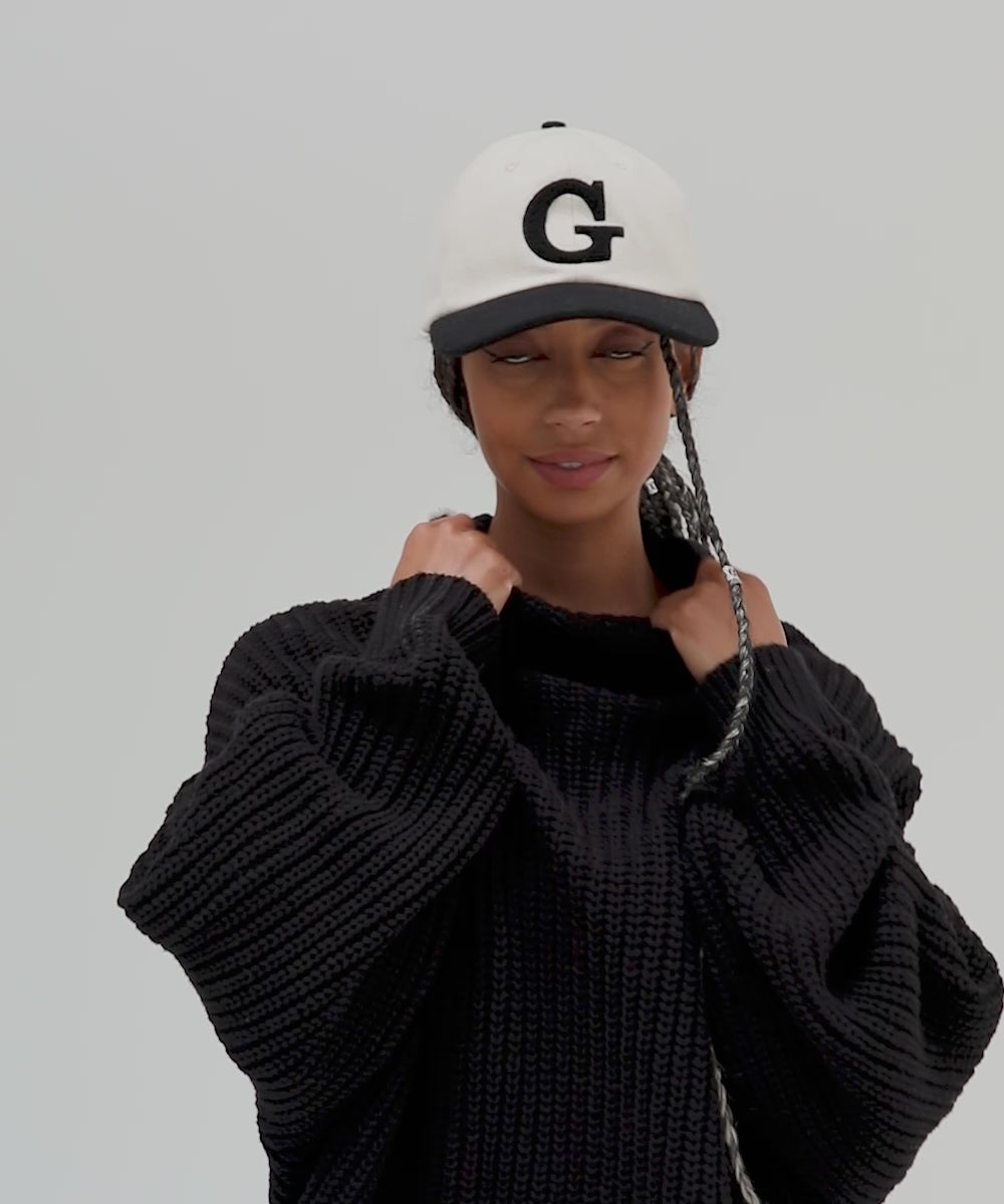 Woman wearing a cream and black baseball cap with the letter 'G' and a black sweater, posing against a plain background #color_cream-black