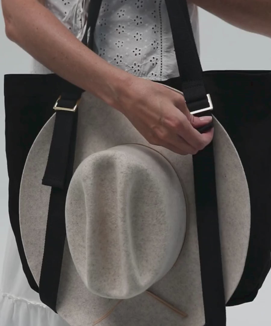 Woman carrying a hat on a black canvas tote bag with two adjustable straps against a plain background #color_black