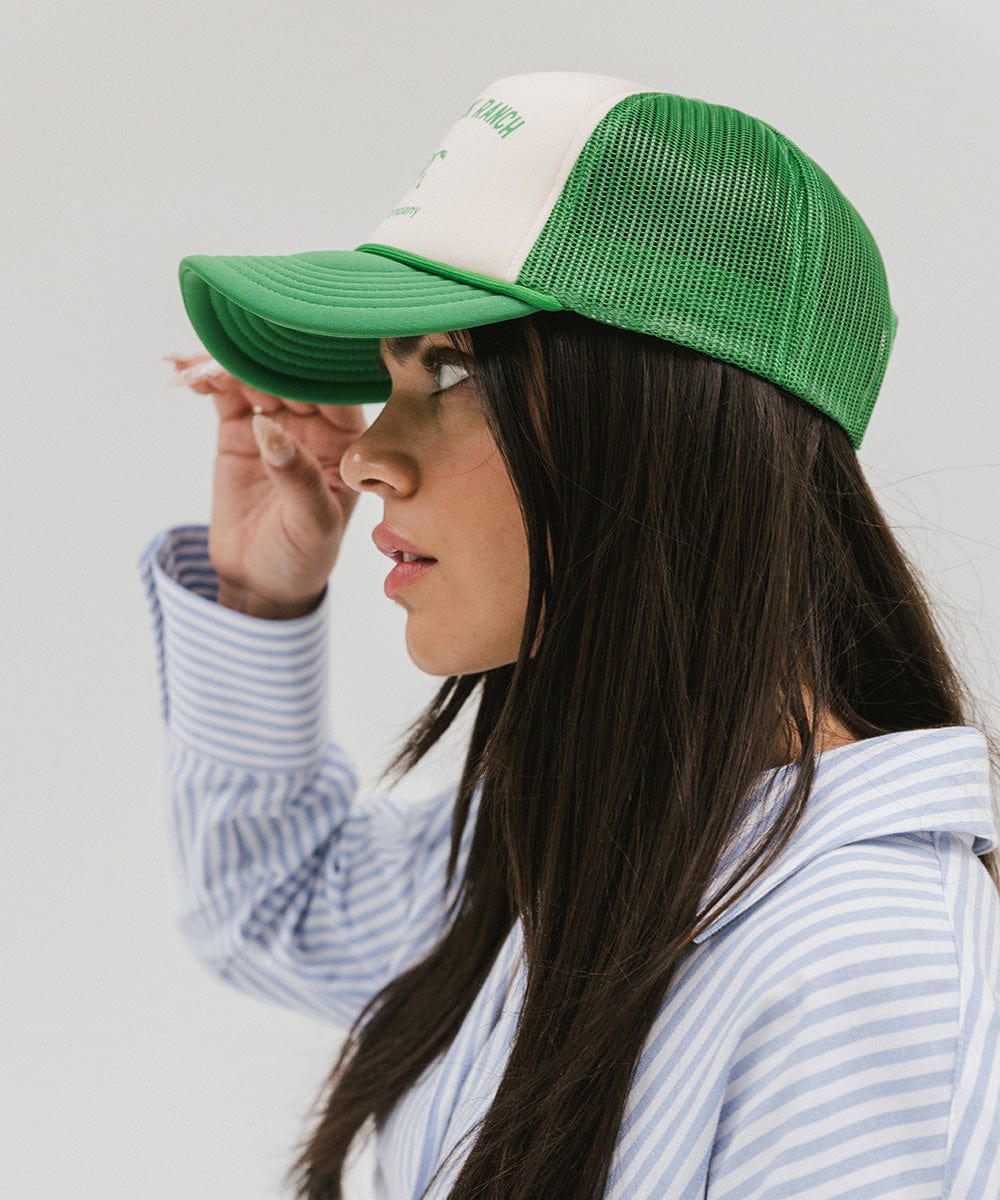 Woman wearing a cream and vintage green cap with 'Saddleback Ranch' text and logo, and a shirt, facing sideways against a plain background #color_cream - vintage green