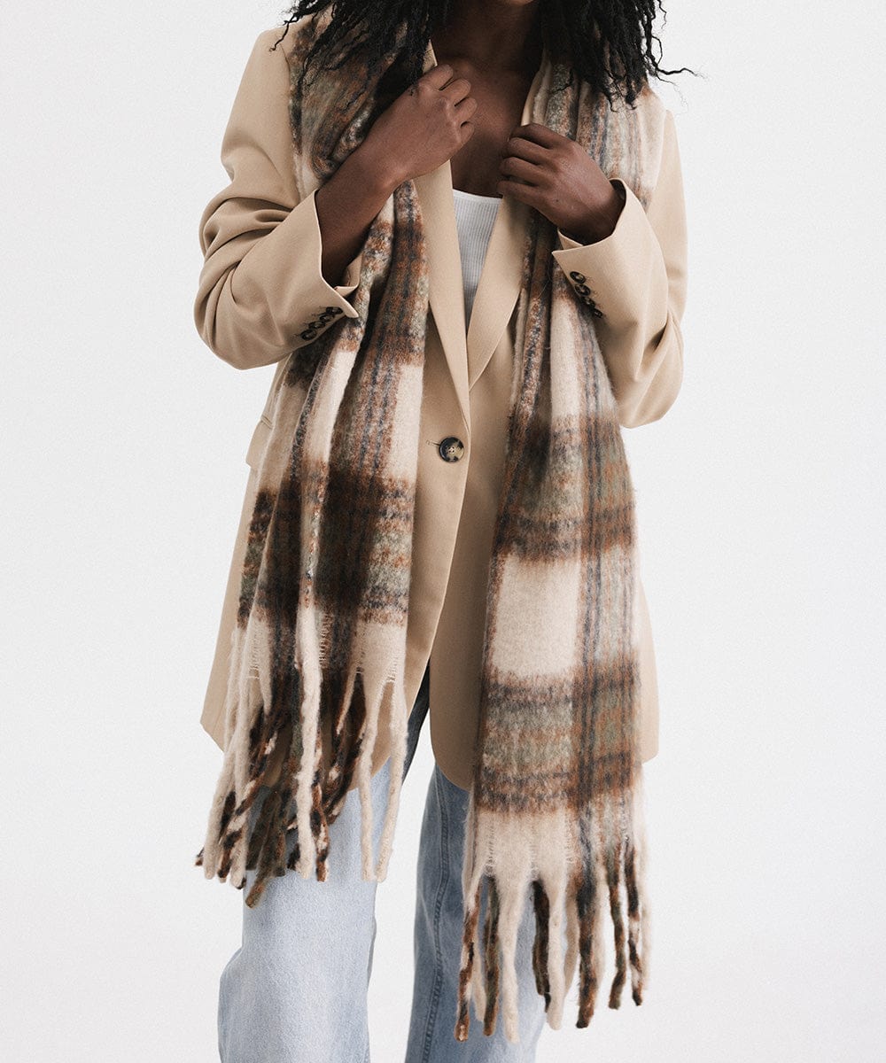 A person wearing a coat and a cream and tan colored oversized knit scarf with fringes, draped over the neck against a plain background #color_cream - tan