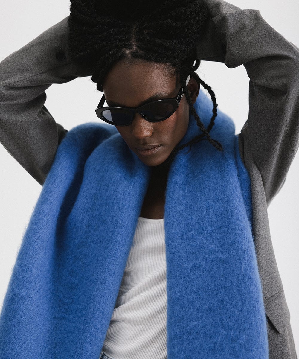 A woman wearing sunglasses, a grey blazer, blue jeans, and an alpine blue colored oversized knit scarf with fringes, draped over the neck against a plain background #color_alpine blue