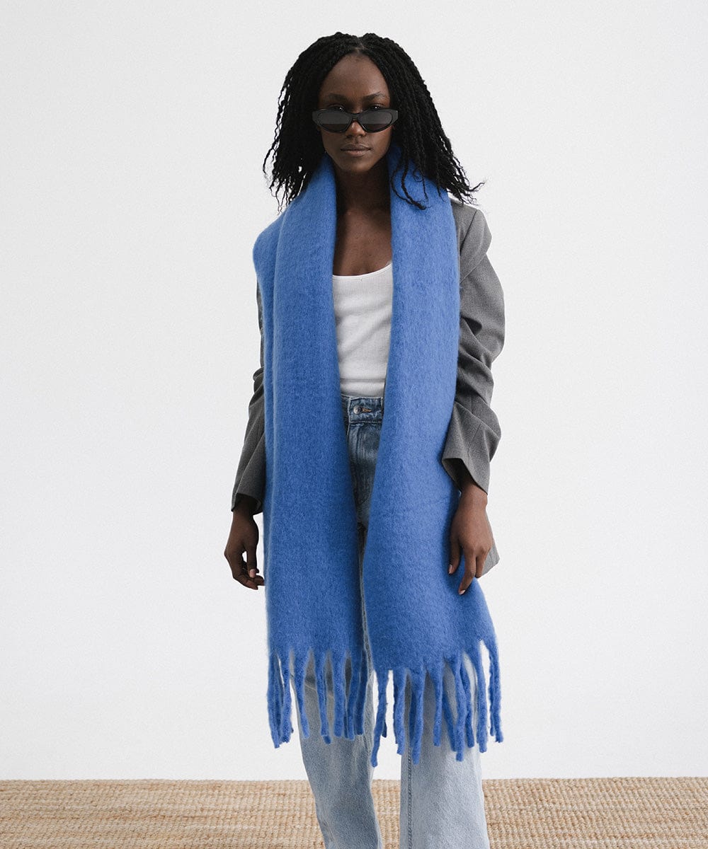 A woman wearing sunglasses, a grey blazer, blue jeans, and an alpine blue colored oversized knit scarf with fringes, draped over the neck against a plain background #color_alpine blue