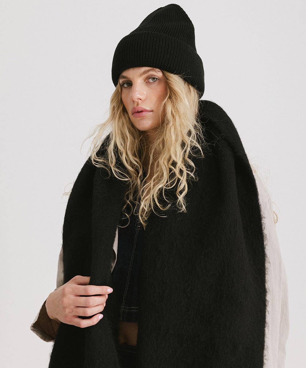 A person wearing a black beanie, and a black oversized knit scarf with fringes, draped over the neck against a plain background #color_black