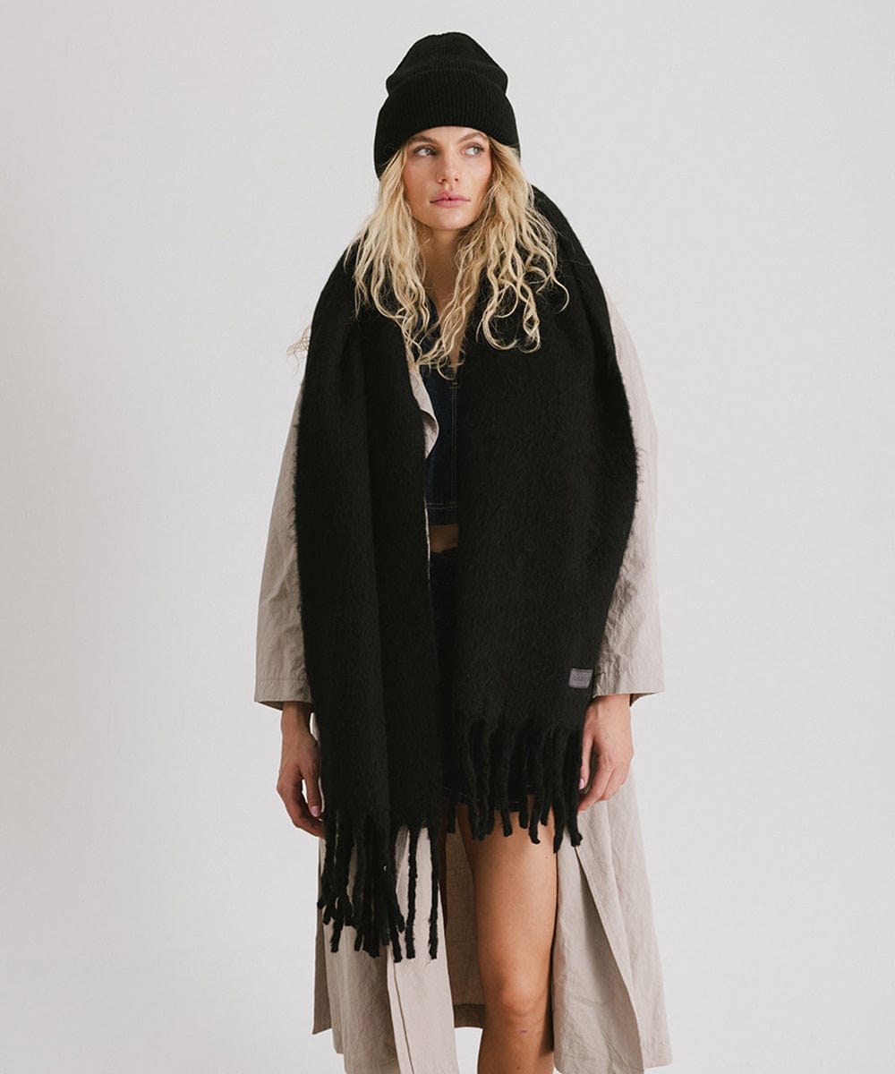 A person wearing a black beanie, a cream coat, and a black oversized knit scarf with fringes, draped over the neck against a plain background #color_black