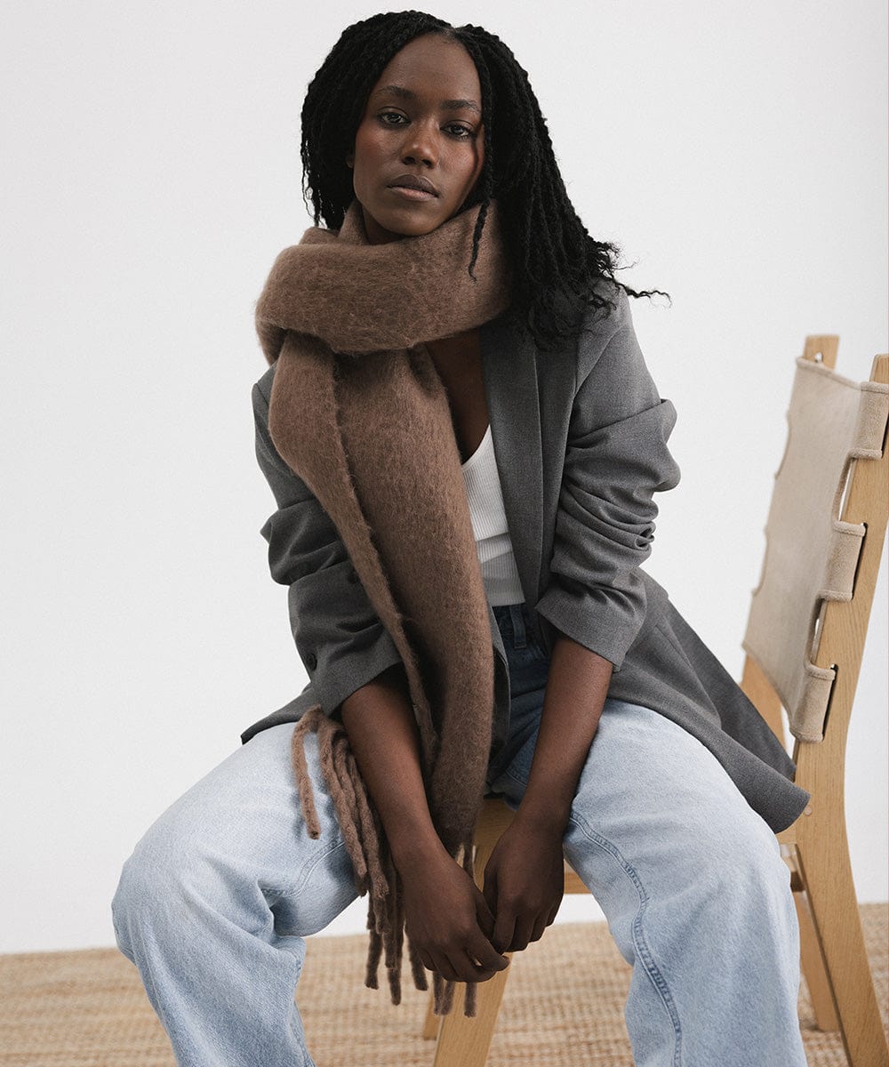A person wearing a grey blazer and blue jeans, and a brown oversized knit scarf with fringes, draped over the neck and sitting on a chair against a plain background #color_brown