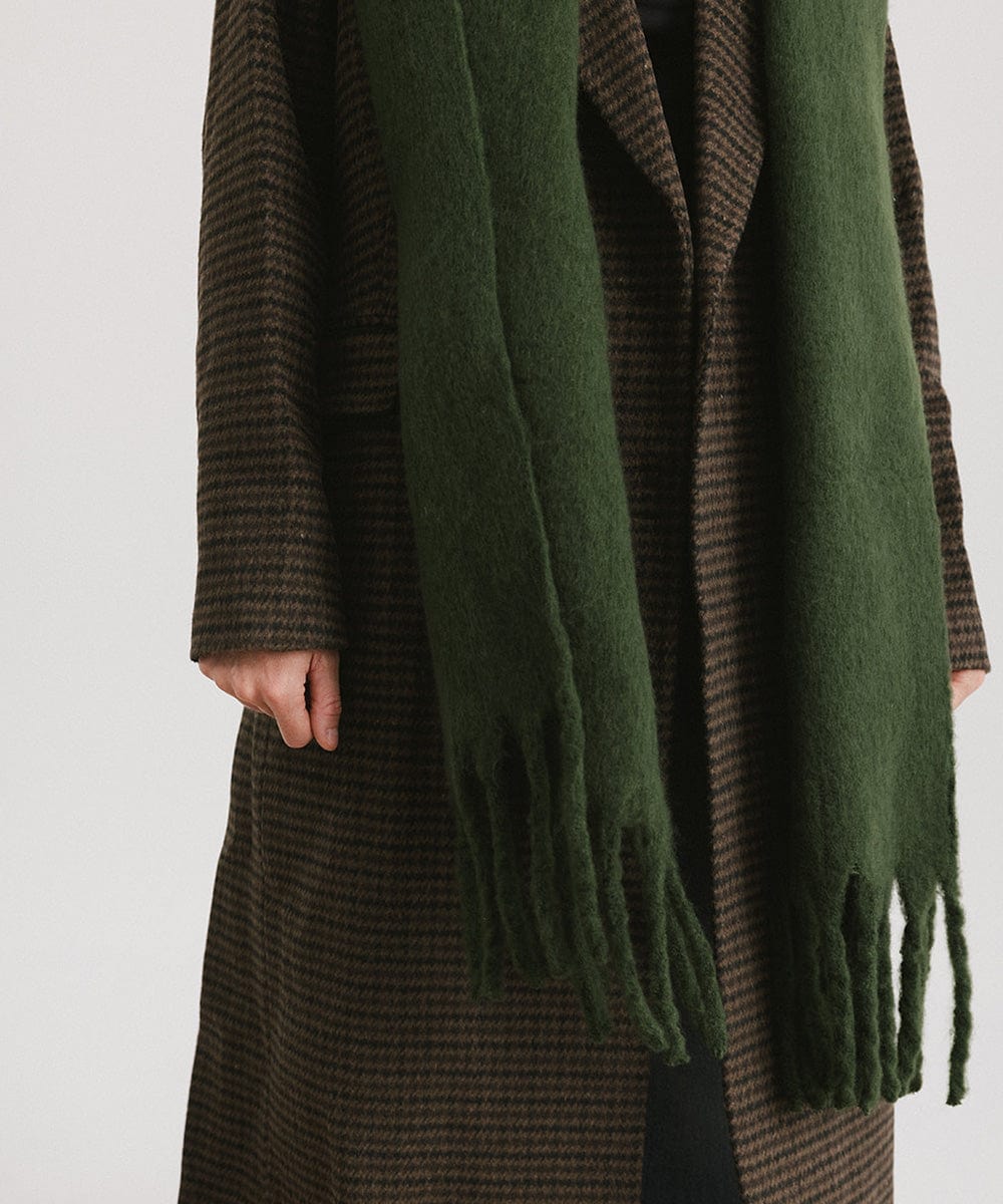 A person wearing a coat and a dark green oversized knit scarf with fringes, draped over the neck against a plain background #color_dark green