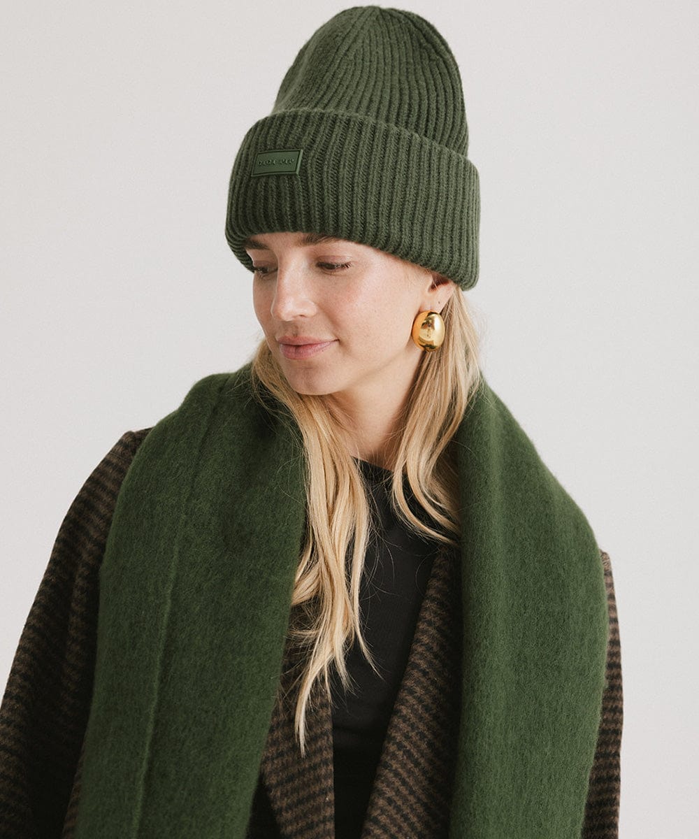 A Woman wearing a dark green beanie, a coat and a dark green oversized knit scarf with fringes, draped over the neck against a plain background #color_dark green