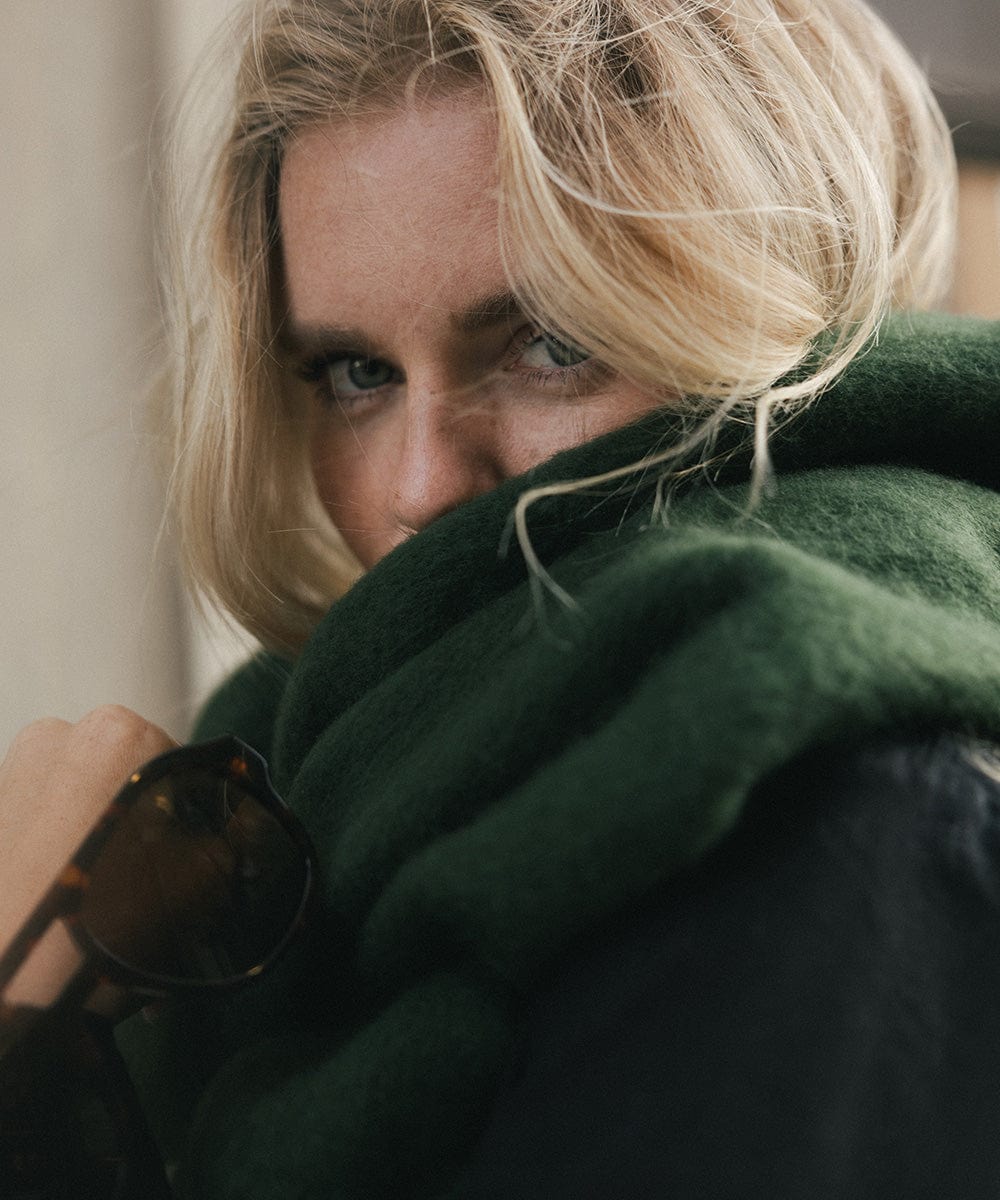 A person wearing a dark green oversized knit scarf with fringes, draped over the neck against a plain background #color_dark green