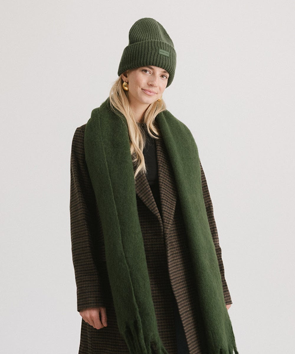 A person wearing a dark green beanie, a coat and a dark green oversized knit scarf with fringes, draped over the neck against a plain background. #color_dark green