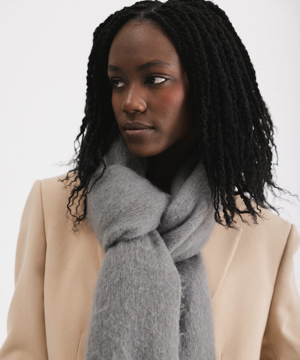 A woman wearing a beige blazer, and a grey oversized knit scarf with fringes, draped over the neck against a plain background #color_grey