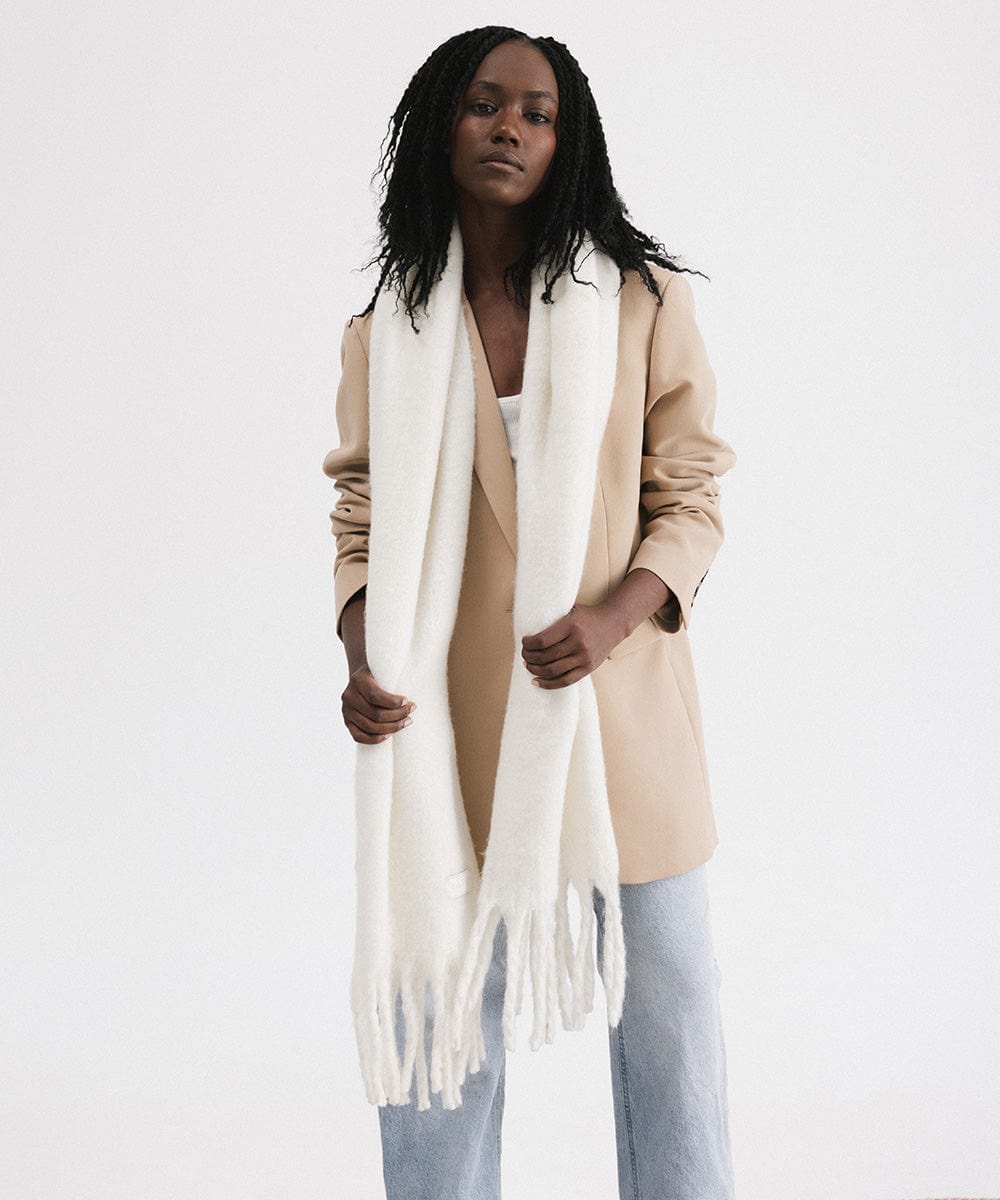 A woman wearing a beige blazer, blue jeans, and an off white oversized knit scarf with fringes, draped over the neck against a plain background #color_off white
