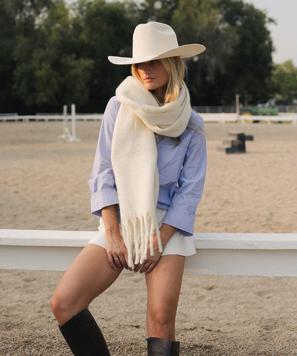 A woman wearing a white hat, blue shirt, white shorts, and an off white oversized knit scarf with fringes, draped over the neck and standing outdoors #color_off white