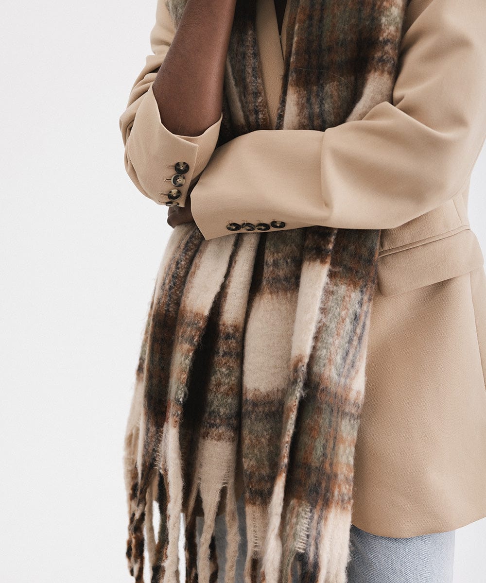 A person wearing a cream and tan colored oversized knit scarf with fringes, draped over the neck against a plain background #color_cream - tan