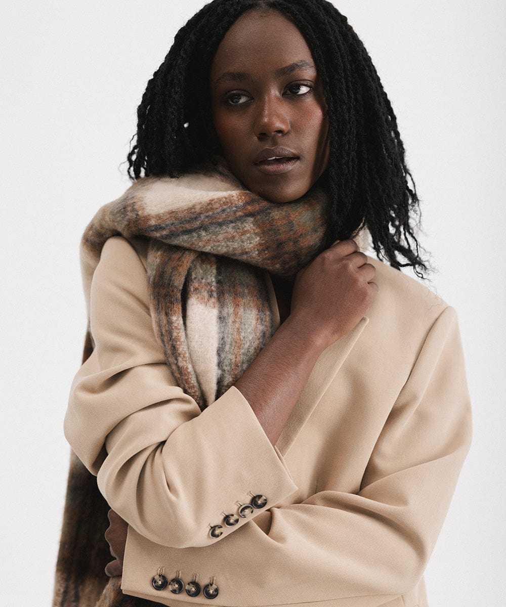A person wearing a coat and a cream and tan colored oversized knit scarf with fringes, draped over the neck against a plain background #color_cream - tan