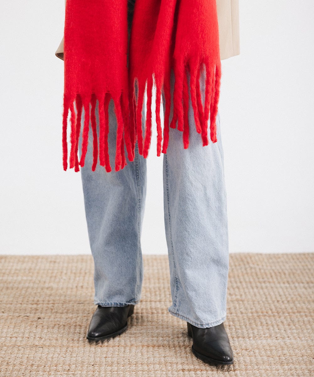 A person wearing a red oversized knit scarf with fringes, and blue jeans with black shoes against a plain background #color_red