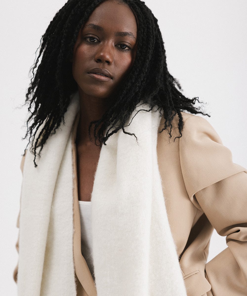 A person wearing a beige blazer and an off white oversized knit scarf with fringes, draped over the neck against a plain background #color_off white