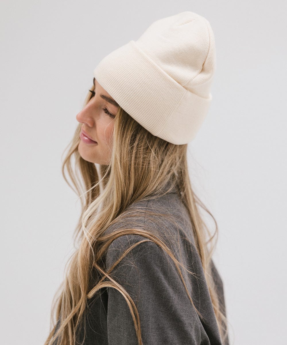 Woman wearing a cream beanie and a grey coat, facing sideways against a plain background #color_cream