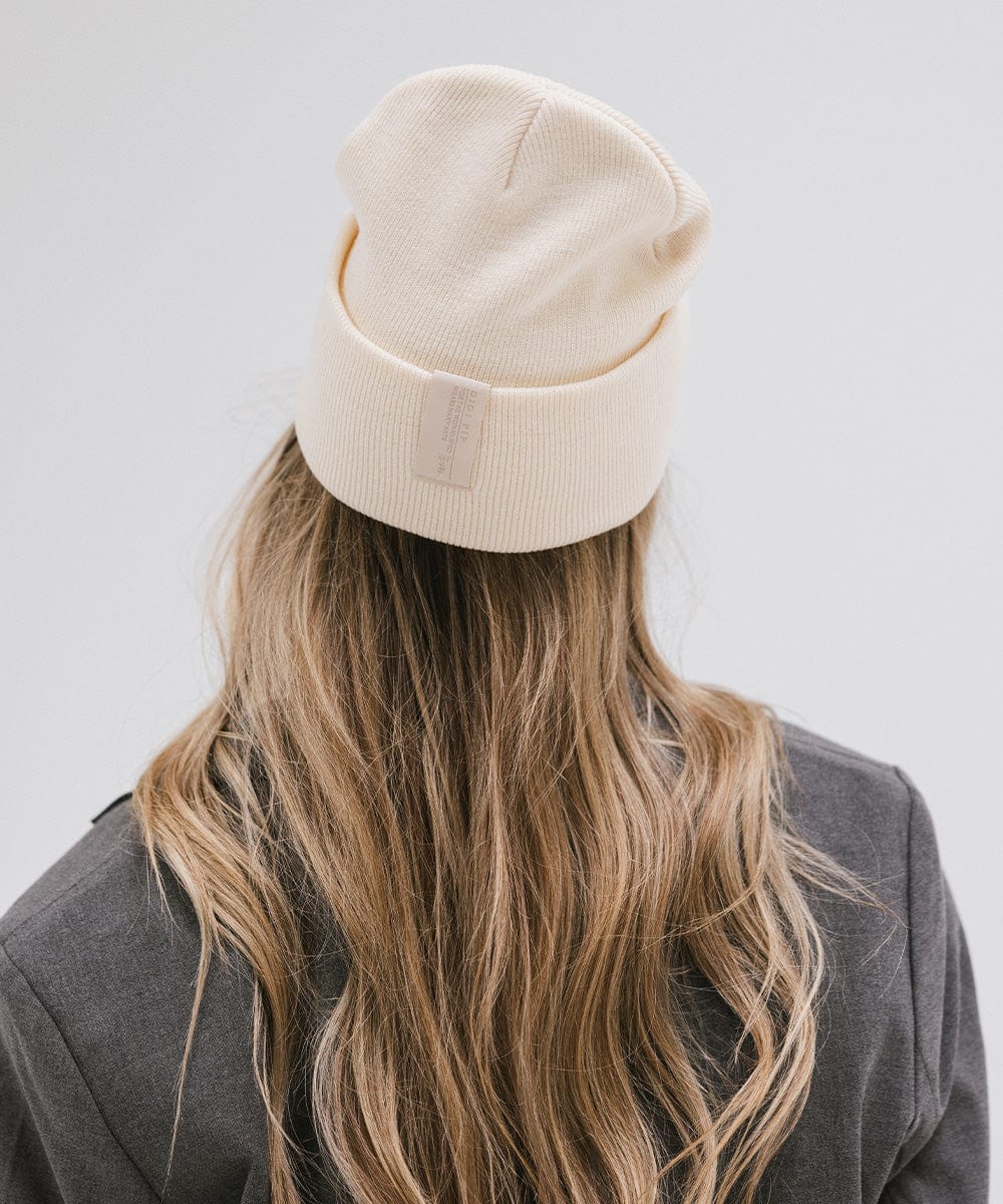 Woman with long hair wearing a cream beanie and a grey coat, facing behind against a plain background #color_cream