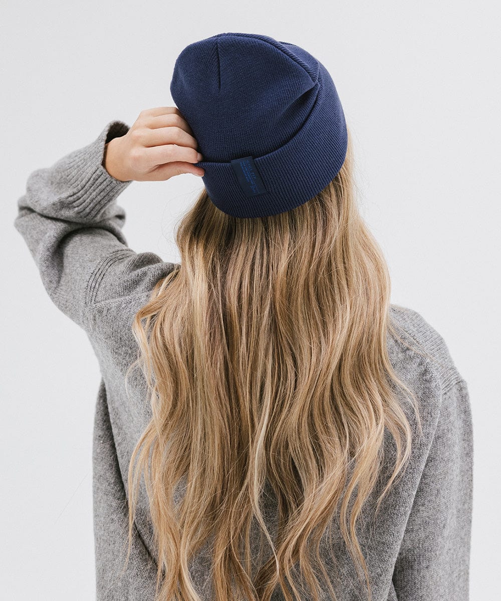 Woman with long hair wearing a navy beanie and a grey sweater, facing behind against a plain background #color_navy