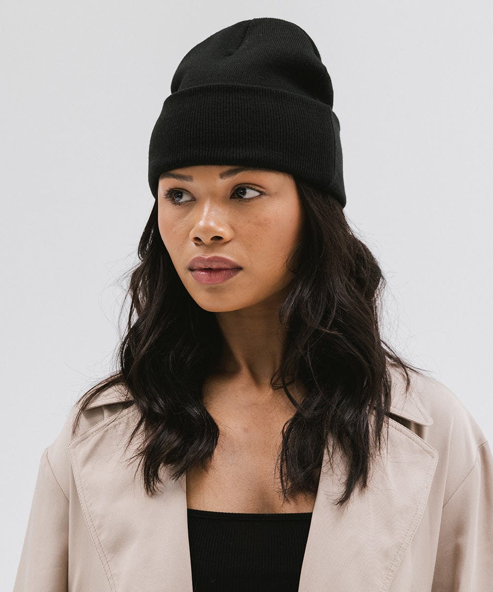 Woman wearing a black beanie and a black top with a cream coat against a plain background #color_black