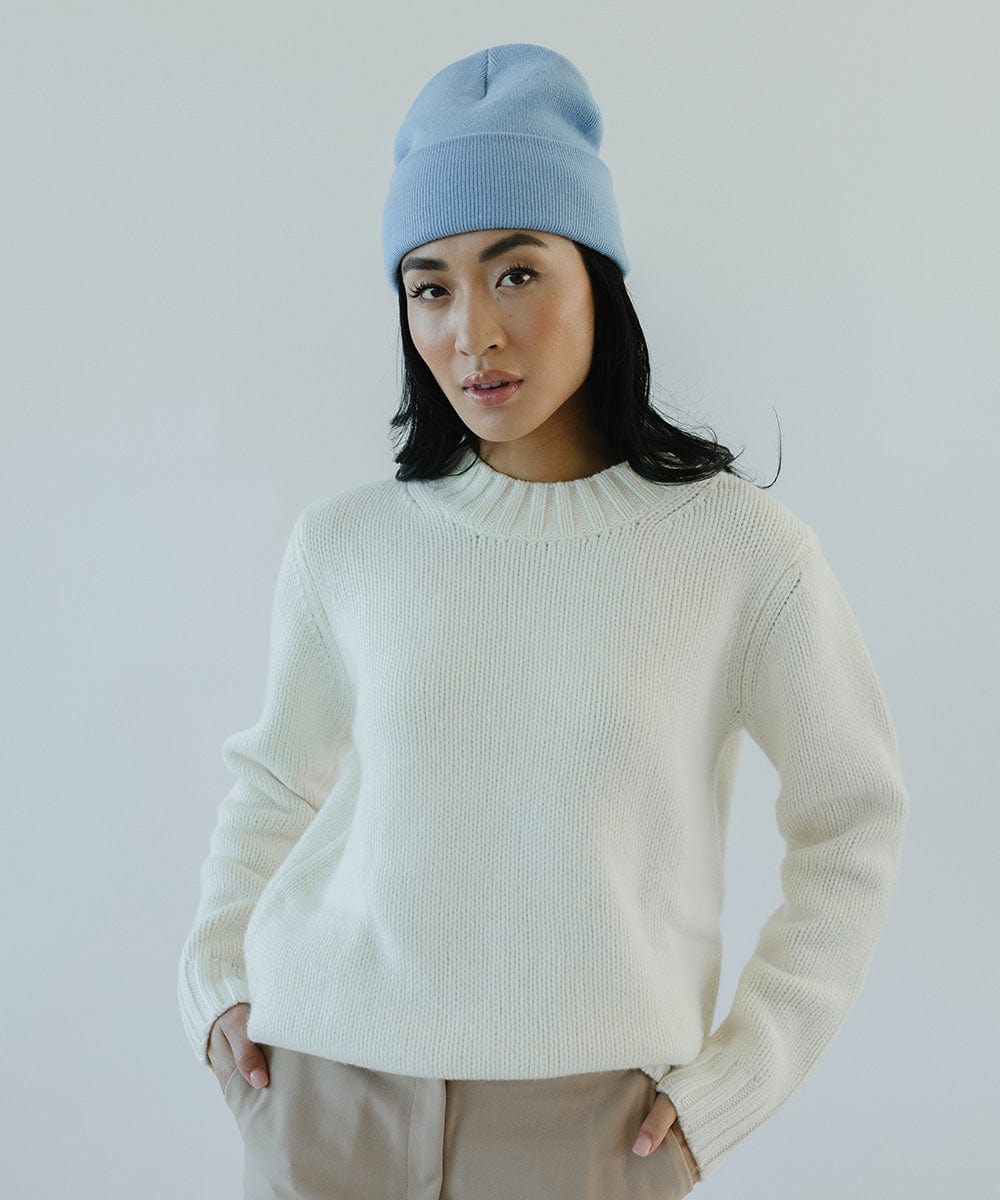 Woman wearing an azure blue colored beanie, a white sweater and beige pants against a plain background #color_azure blue