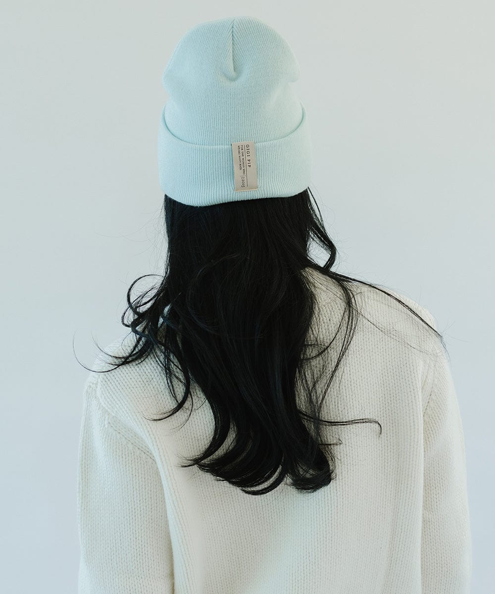 Woman with long hair wearing a sky blue beanie with the brand label and a white sweater, facing behind against a plain background #color_sky blue