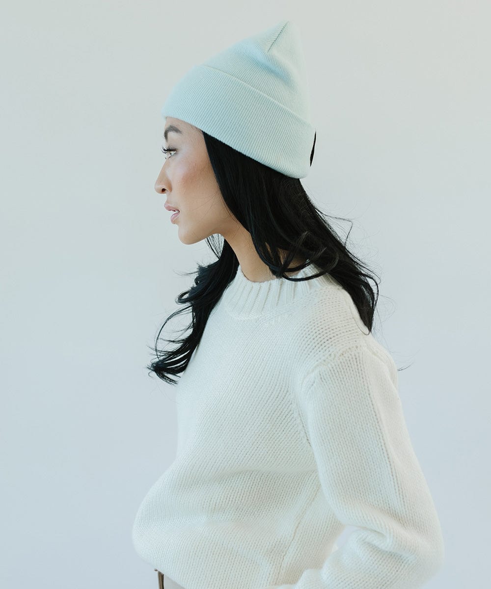 Woman wearing a sky blue beanie and a white sweater, facing sideways against a plain background #color_sky blue