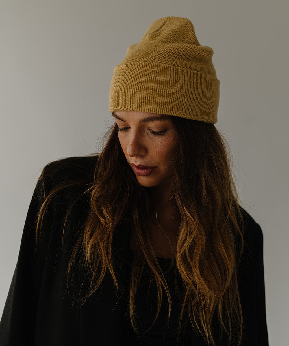 Woman wearing a honeycomb colored beanie and a black coat, looking down against a plain background #color_honeycomb
