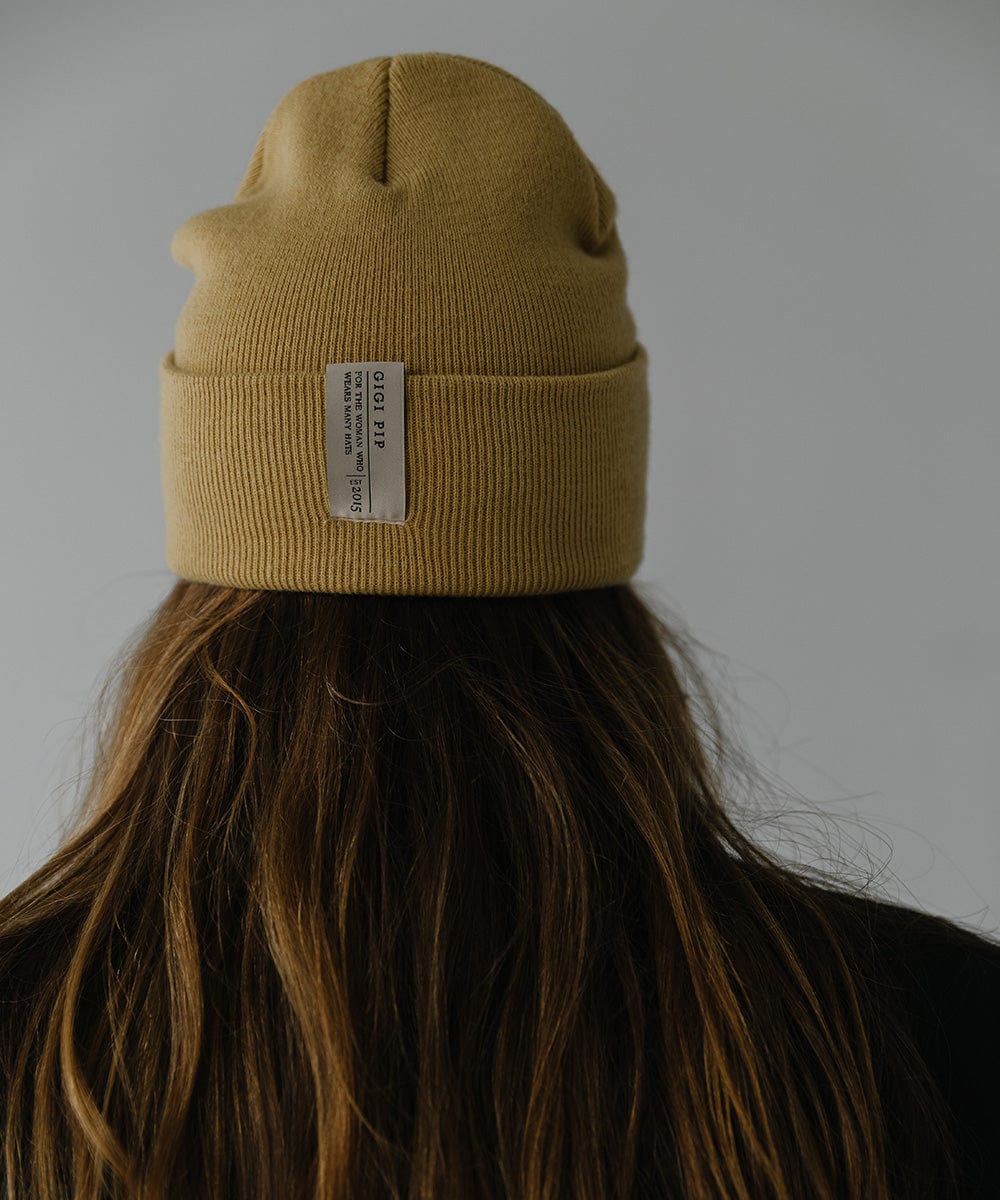 Woman wearing a honeycomb colored beanie with the brand label and a black coat, facing behind against a plain background #color_honeycomb