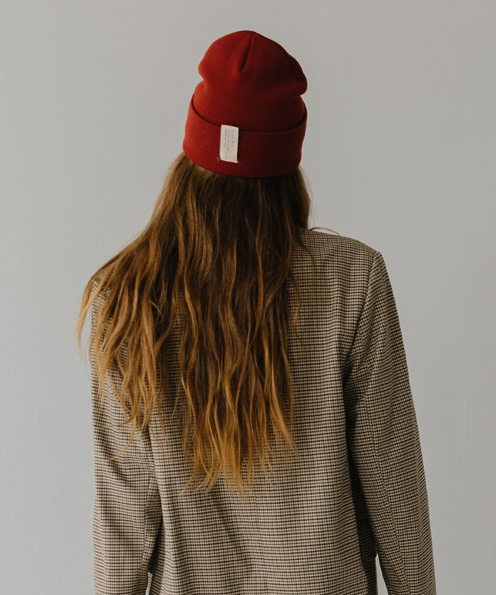 Woman with long hair wearing a brick red colored beanie and a coat, facing behind against a plain background #color_brick red