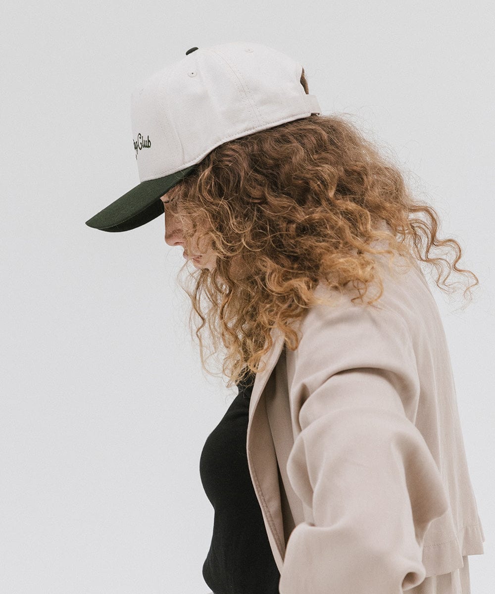 Woman wearing a cream and dark green cap with 'Slow Morning Club' text and a black top with a beige coat, facing sideways against a plain background #color_cream - dark green