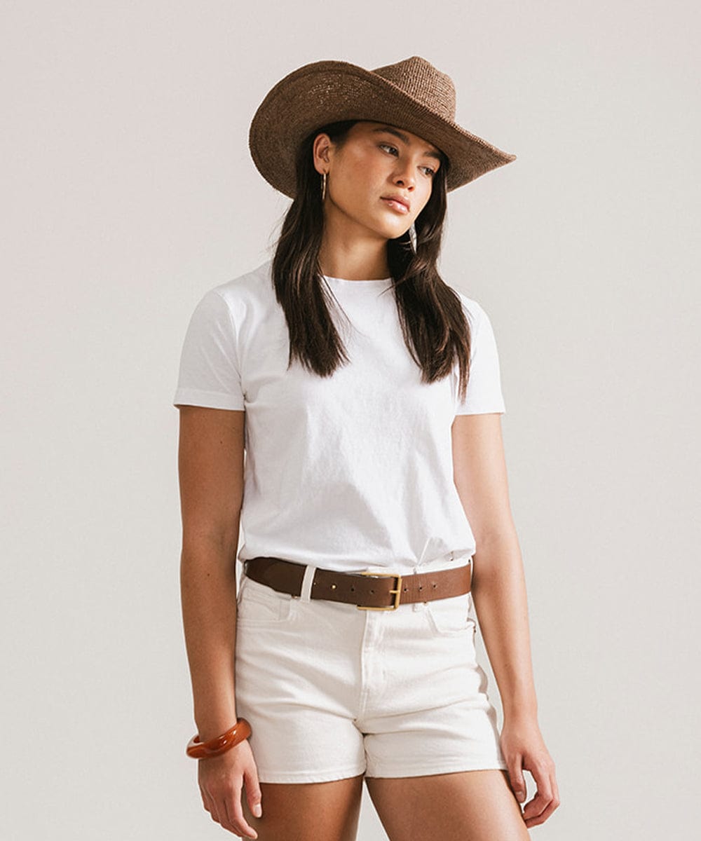 Woman wearing a chocolate colored straw cowboy hat, a white t-shirt, brown belt and white shorts against a plain background #color_chocolate