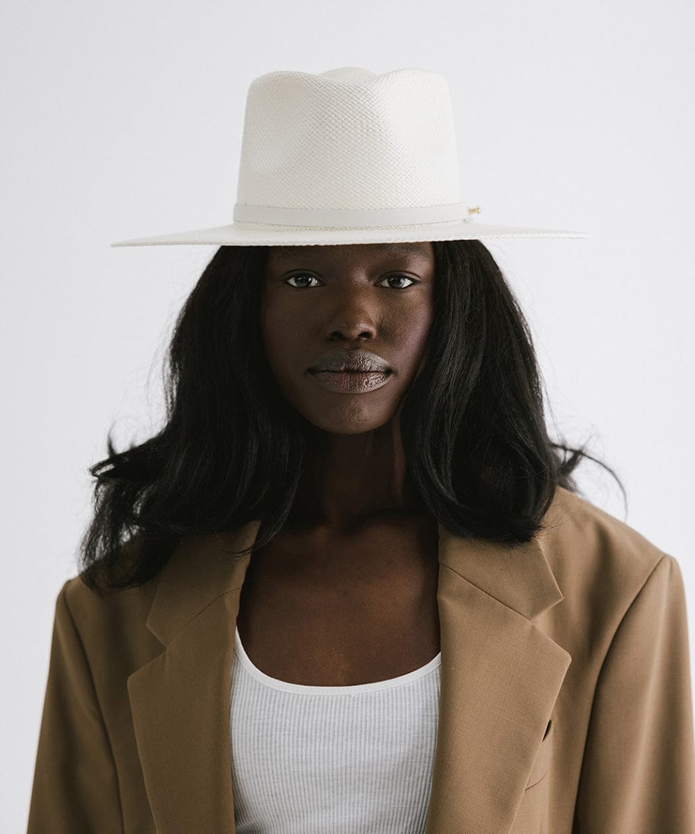 Woman wearing a white straw hat with a hat band and a brown coat against a plain background #color_white