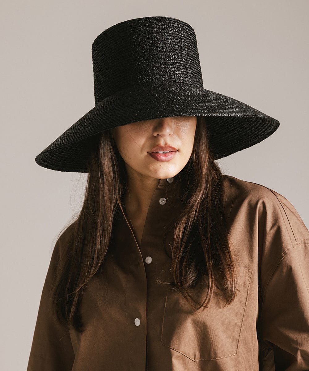 Woman wearing a black oversized raffia straw bucket hat and a brown shirt against a plain background #color_black