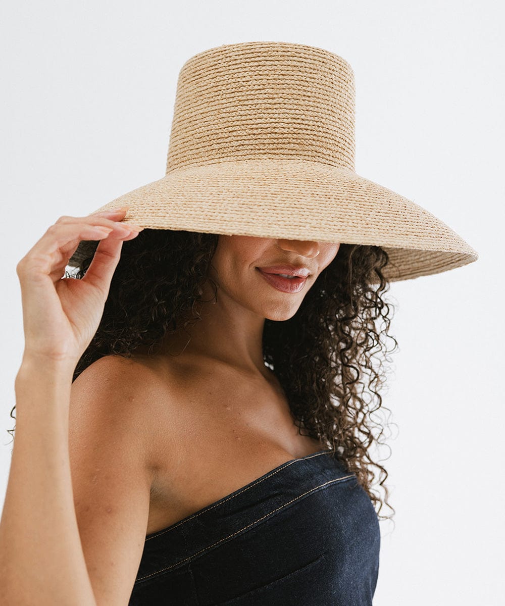 Woman with curly hair wearing a natural colored oversized raffia straw bucket hat and a denim top against a plain background #color_natural