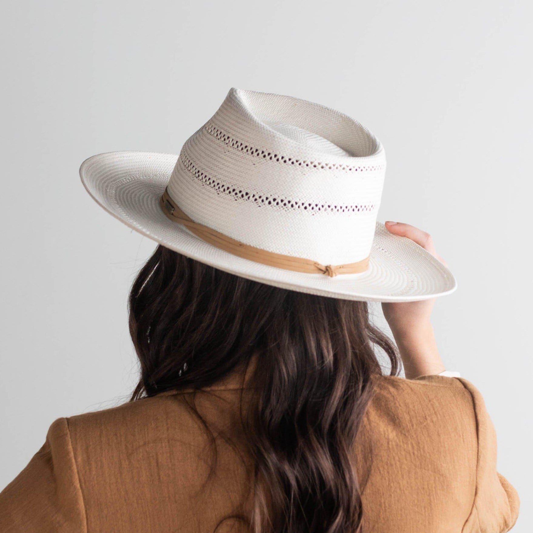 Person wearing a tan straw fedora hat with a hat band and a brown coat against a plain background #color_tan