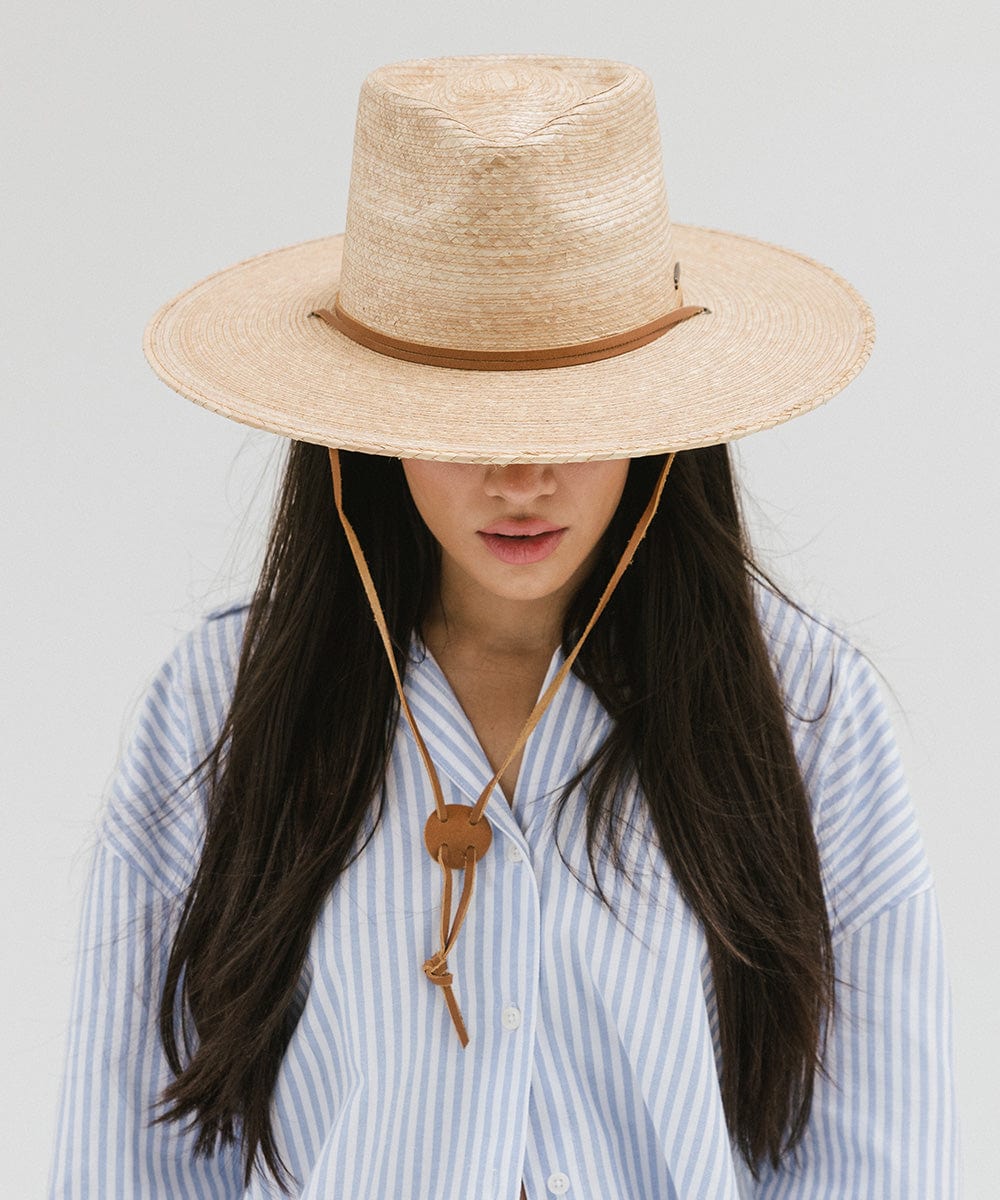 Woman wearing an oak colored palm straw sun hat with a brown chinstrap and a white shirt with blue lines, looking down against a plain background #color_oak
