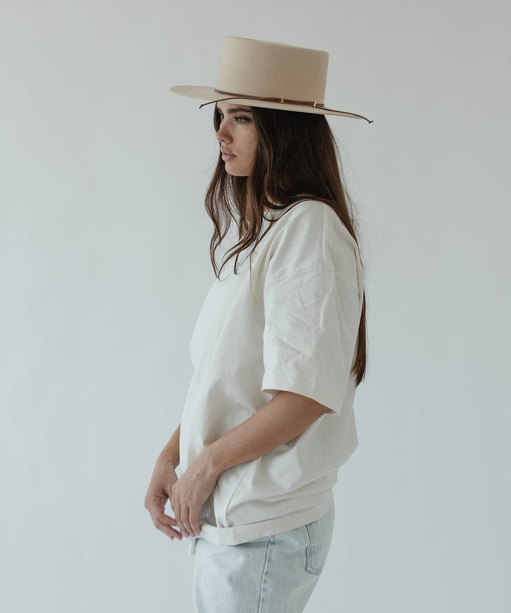 Woman wearing a beige hat with a brown suede band, a white t-shirt and light blue jeans, facing sideways against a plain background #color_brown