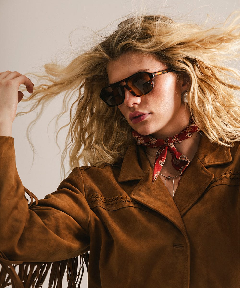 Woman wearing sunglasses and a brown coat with a red scarf, against a neutral background #color_tortoise